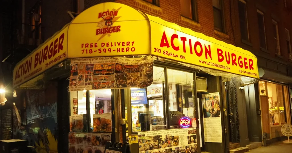At night, Action Burger in NYC glows under a yellow awning, with comic posters and a neon OPEN sign in the window.
