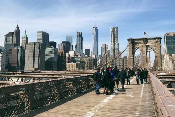 People walk on Brooklyn Bridge under clear skies; New York City skyline and iconic cables prominently displayed.