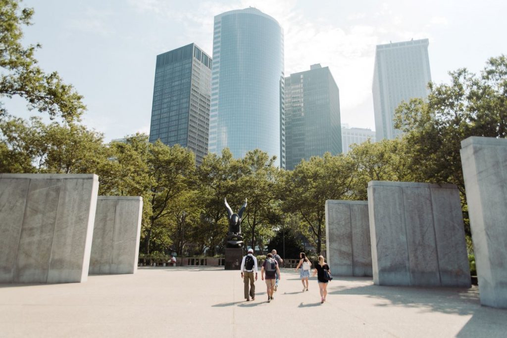 People stroll in a plaza surrounded by skyscrapers and trees, with a sculpture centerpiece under partly cloudy skies.