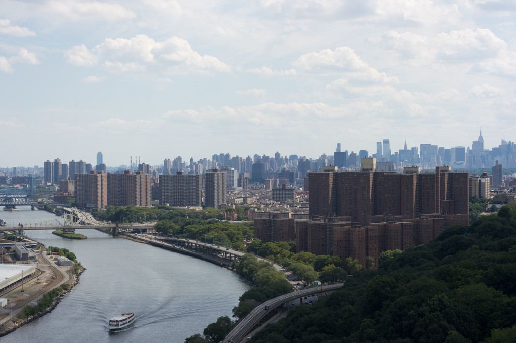 A boat travels down the Harlem River in New York City, passing green trees and tall apartments under a partly cloudy sky.