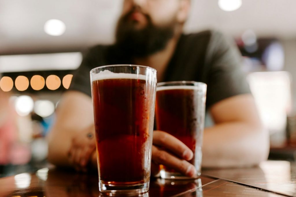 Two beers on a table and a man in the background sitting with his beer at markets in NYC. 