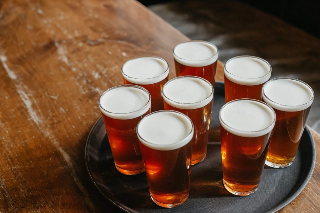Eight frothy pints of amber beer sit on a round black tray atop a rustic wooden pub table, evoking English pub tradition.