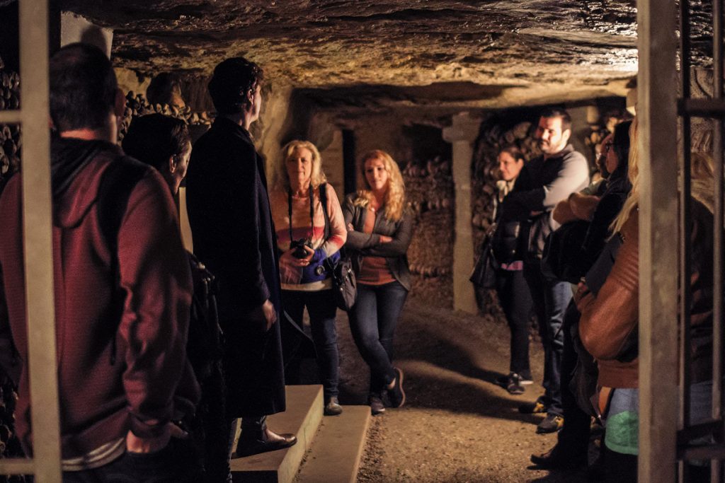 Several people gather around a tour guide in the dimly lit Paris Catacombs, surrounded by neatly stacked skulls and bones.