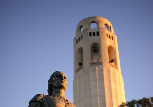 A bronze statue of Christopher Columbus stands before Coit Tower in San Francisco, surrounded by trees at sunset.