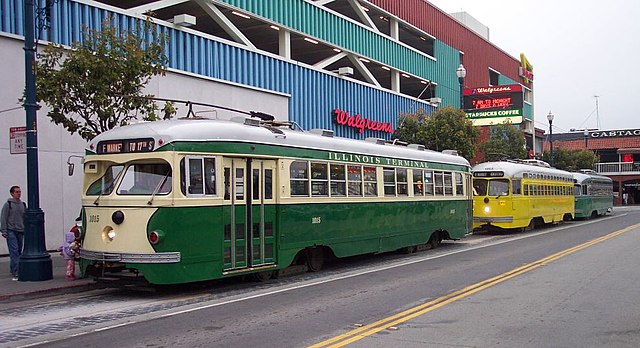 Two vintage streetcars, including a green and cream Illinois Terminal, are parked on Market Street in San Francisco as people gather nearby.