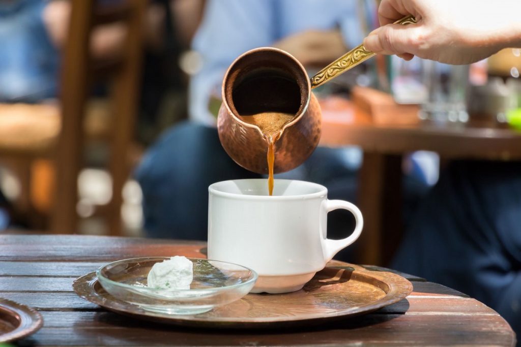 Hand pouring coffee from a traditional copper pot into a white coffee mug