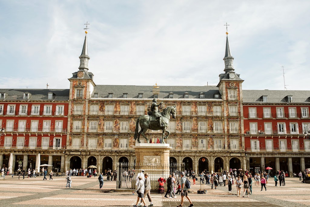 A bustling Plaza Mayor in Madrid features the King Philip III equestrian statue, surrounded by vibrant crowds and ornate red-and-white architecture.
