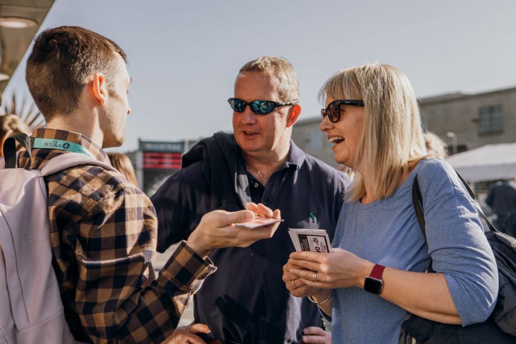 Three people converse outdoors in bright sunlight; a young man gestures while speaking to a man and woman wearing sunglasses.
