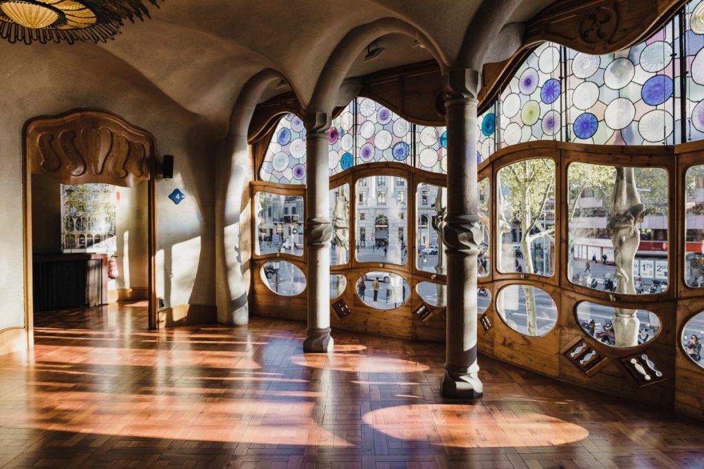 Inside Casa Batlló showing beautiful stained glass windows, curved ceilings, and tree-like pillars. Sunlight pours in through the windows, offering a glimpse of Barcelona's prominent street, Passeig de Gràcia.
