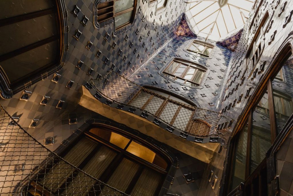 A view from the bottom looking up to the ceiling of an interior chamber of Casa Batlló, one of Antoni Gaudí's famous landmarks. The view shows light cascading through the glass ceiling, highlighting the intricate tile design along the wall and the chamber's many windows.