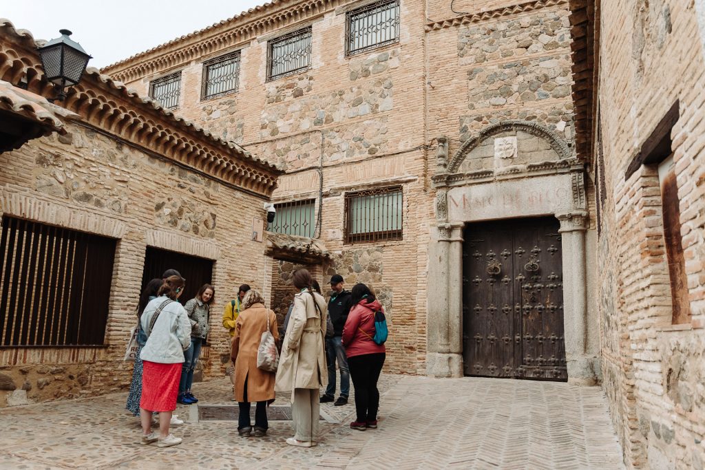 A tour group listens to a guide in a historic European courtyard beside an ornate wooden door and barred stone windows.