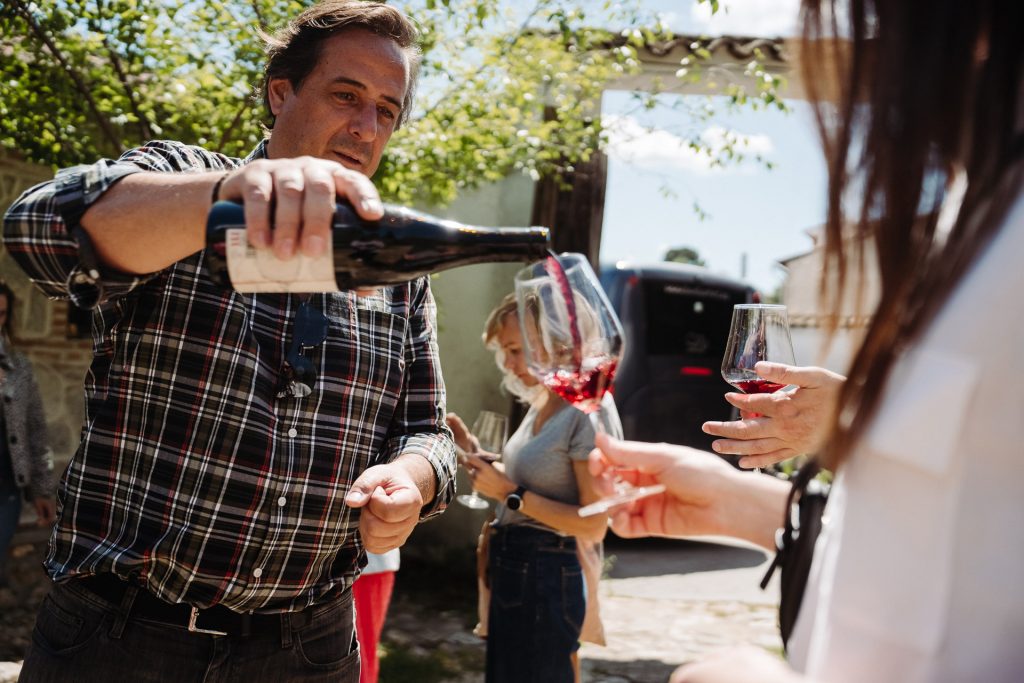 A man in a plaid shirt pours red wine for another person at a Toledo winery, surrounded by trees, friends, and parked cars.