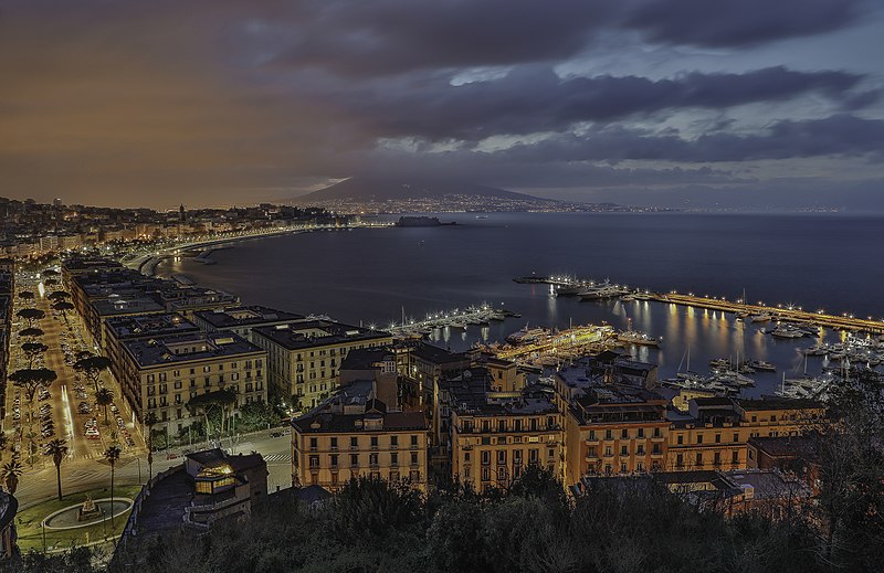 City lights illuminate Naples, Italy’s coastline and marina at dusk, with boats moored and Mount Vesuvius beneath dramatic clouds.
