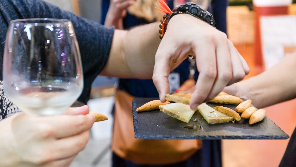A man holds a glass of white wine as another person takes cheese from a slate platter with assorted cheeses and breadsticks. njoy wine and manchego cheese as part of a Toledo Day Trip and Winery Tour.