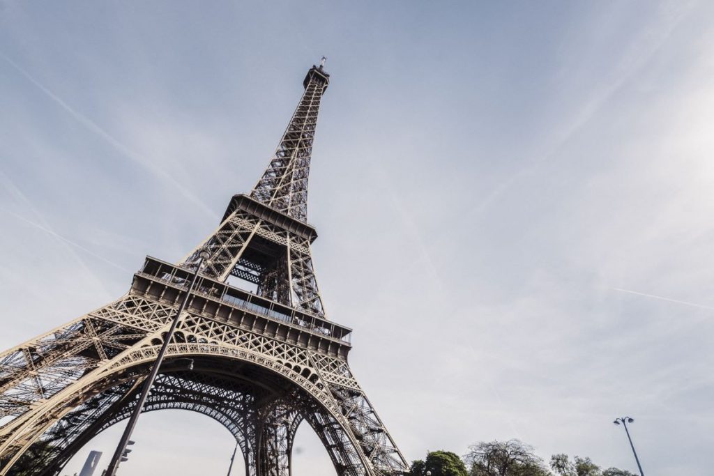 The Eiffel Tower rises above trees and lampposts in Paris, viewed from below against a clear sky with scattered clouds.
