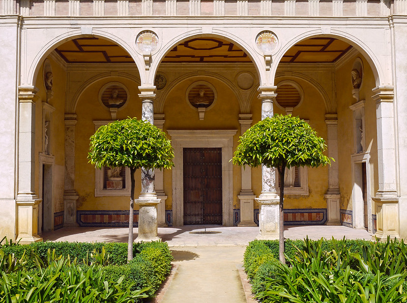 A view of Casa de Pilatos in Seville displaying its unique architectural style, plus a pair of trees and a garden.