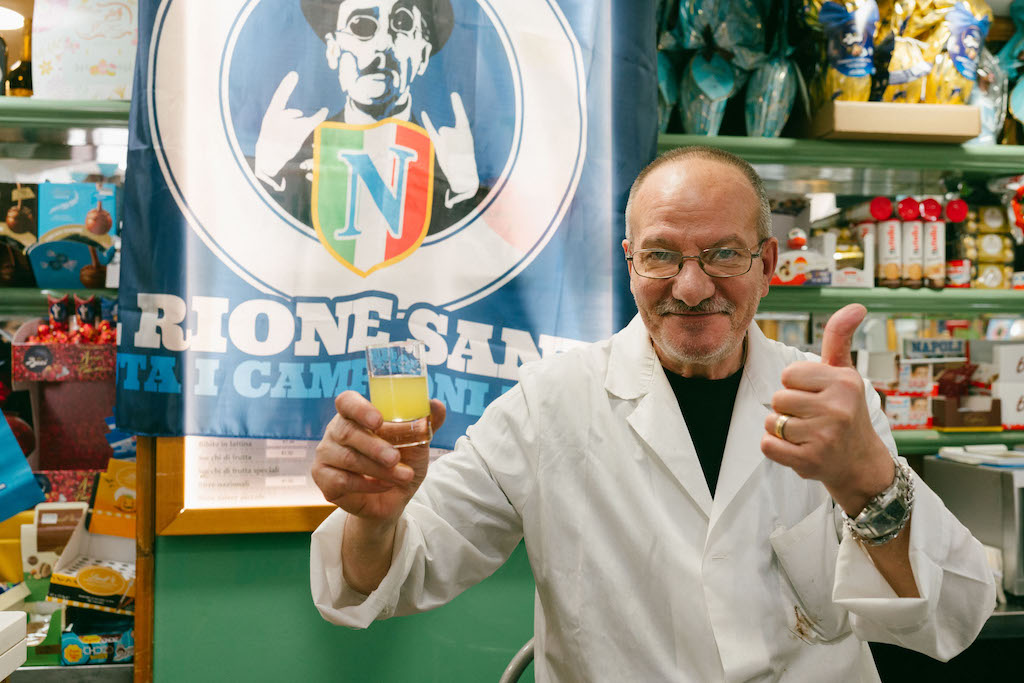 Store owner wearing lab coat-esque outfit holds up a shot of lemoncello in front of a blue flag that reads "Rione Sanità"