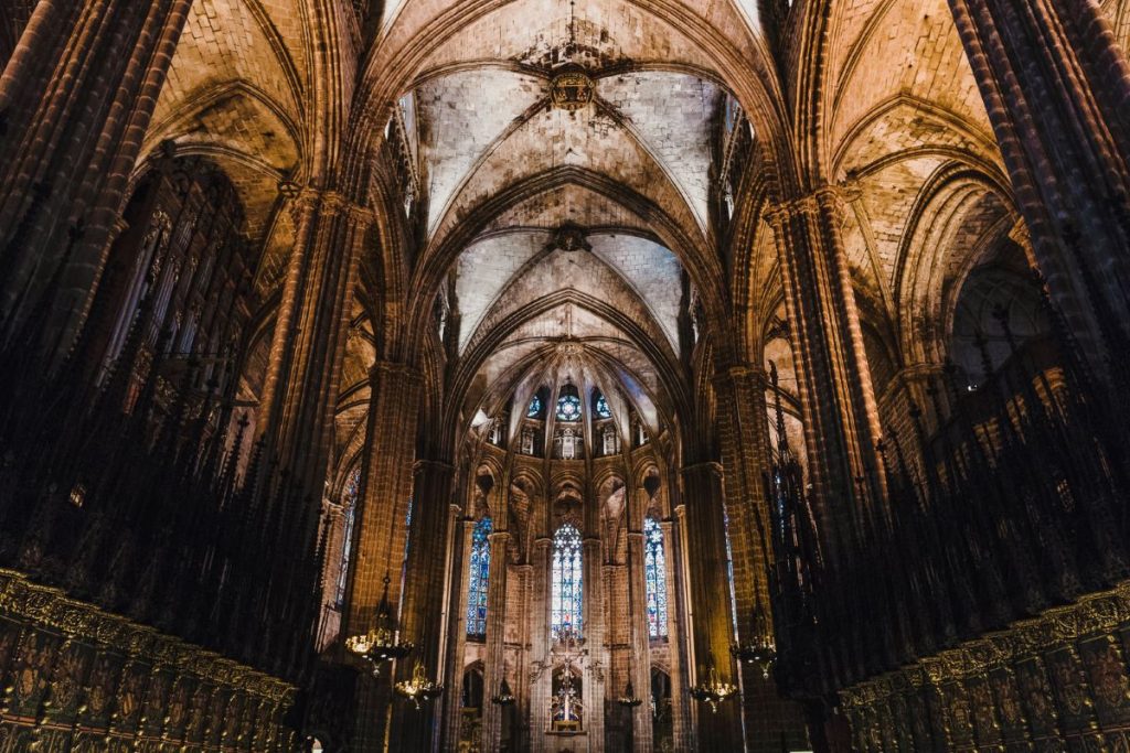 Sunlight streams through tall stained glass windows, illuminating stone arches and ornate details inside the grand vaulted nave of a Gothic cathedral.