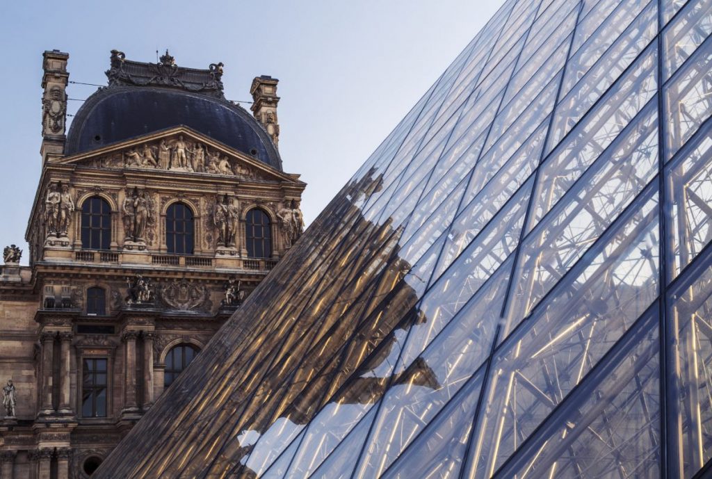 The glass pyramid at the Louvre Museum in Paris reflects ornate sculptures and historic architecture under a bright, clear sky.