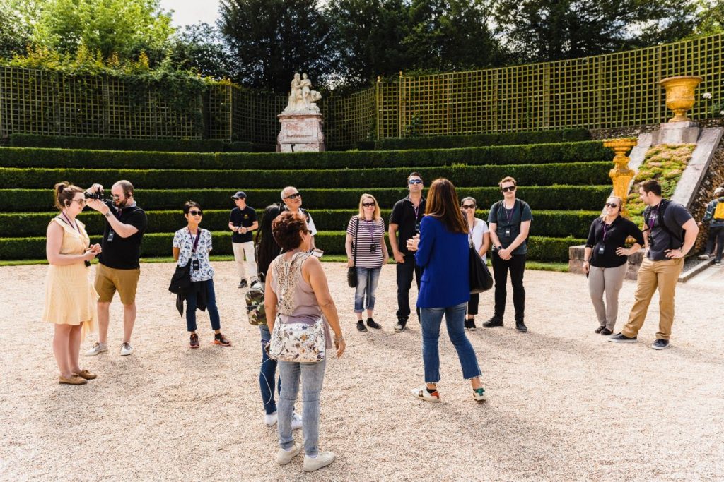 A tour guide speaks to casually dressed visitors in a circular garden with green hedges, trellises, and classical statues, under sunshine.