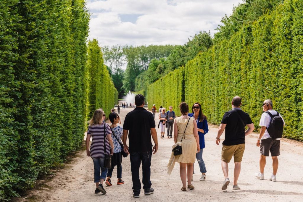 Visitors stroll along a wide gravel path bordered by manicured green hedges in the gardens of Versailles under a partly cloudy sky.