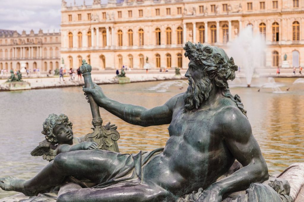 A bronze statue of a reclining bearded man with a staff and cherub rests by a pond at the Palace of Versailles.