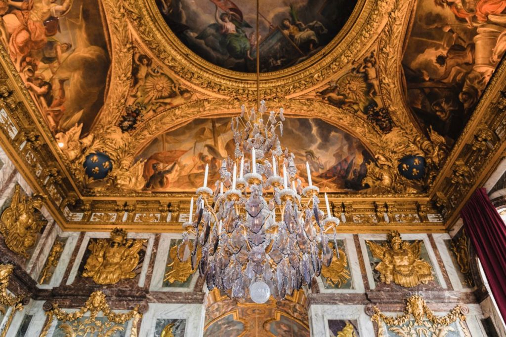 A sparkling chandelier hangs from a gold-accented ceiling adorned with detailed moldings and classical painted murals in Versailles.