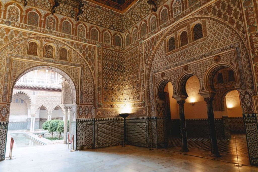 A richly decorated Moorish hall in Alhambra, Granada features intricate tilework, arches, warm lighting, and a courtyard pool.