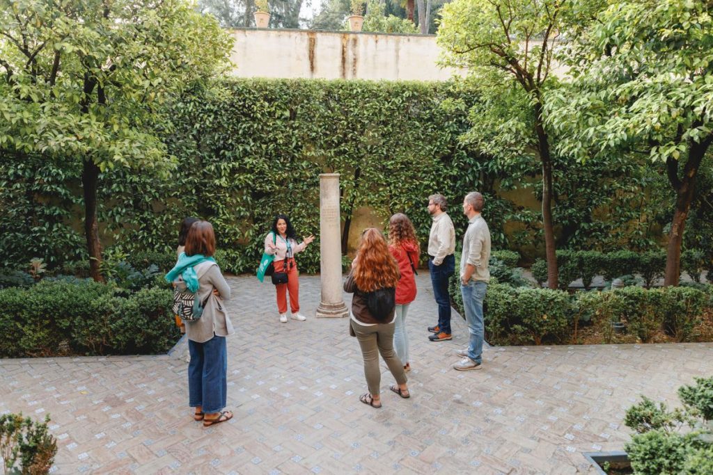 Six people listen to a woman giving a tour in a lush garden courtyard, gathered around a central stone column.