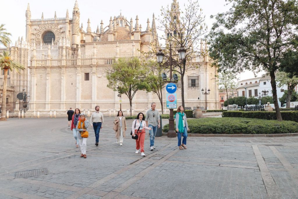 Seven people walk together on a cobblestone plaza in front of an ornate historic cathedral, surrounded by trees and lampposts.