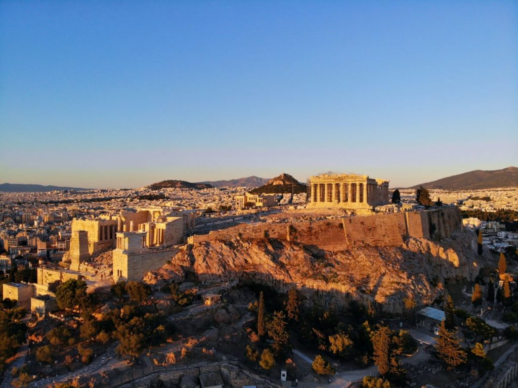 The sun sets over the Acropolis, offering a stunning view. In the background, a large part of the city of Athens is seen.