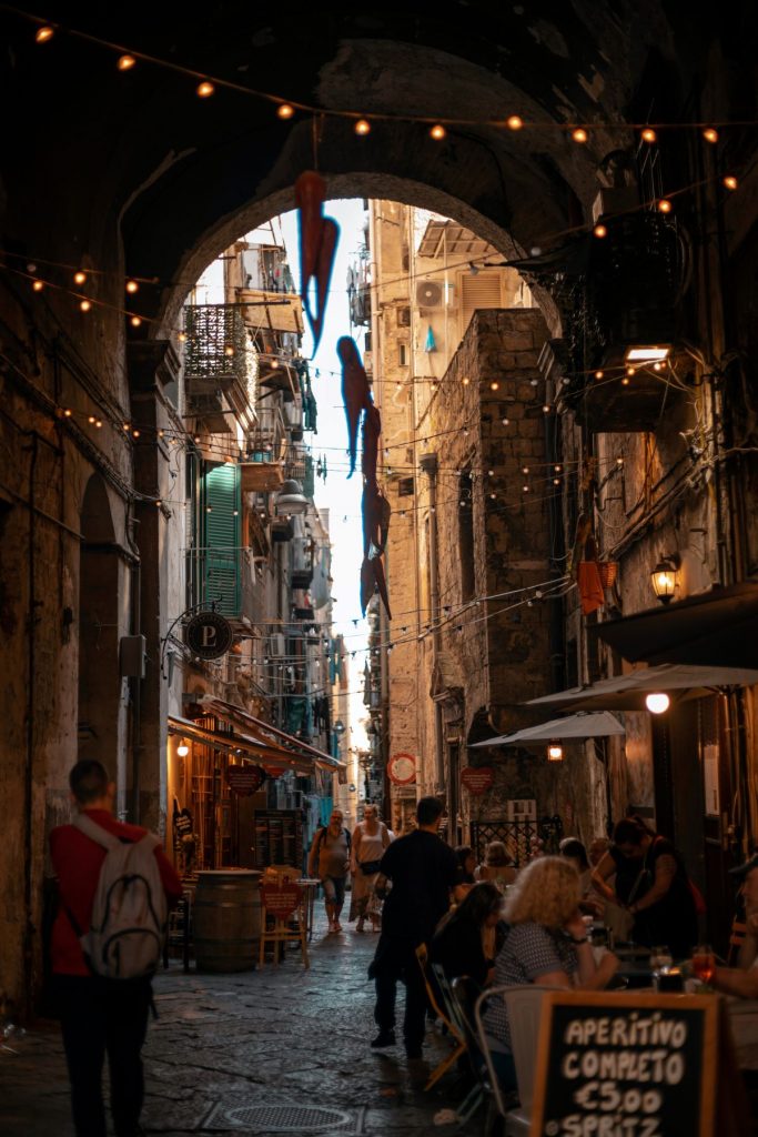 A small street in Naples, Italy, at night filled with people dining or strolling. String lights connected between balconies give it a charming nighttime feeling.