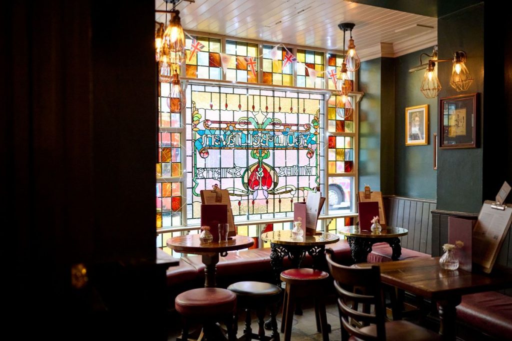 The corner of a pub displaying wooden table and chairs and a beautiful stained glass window.