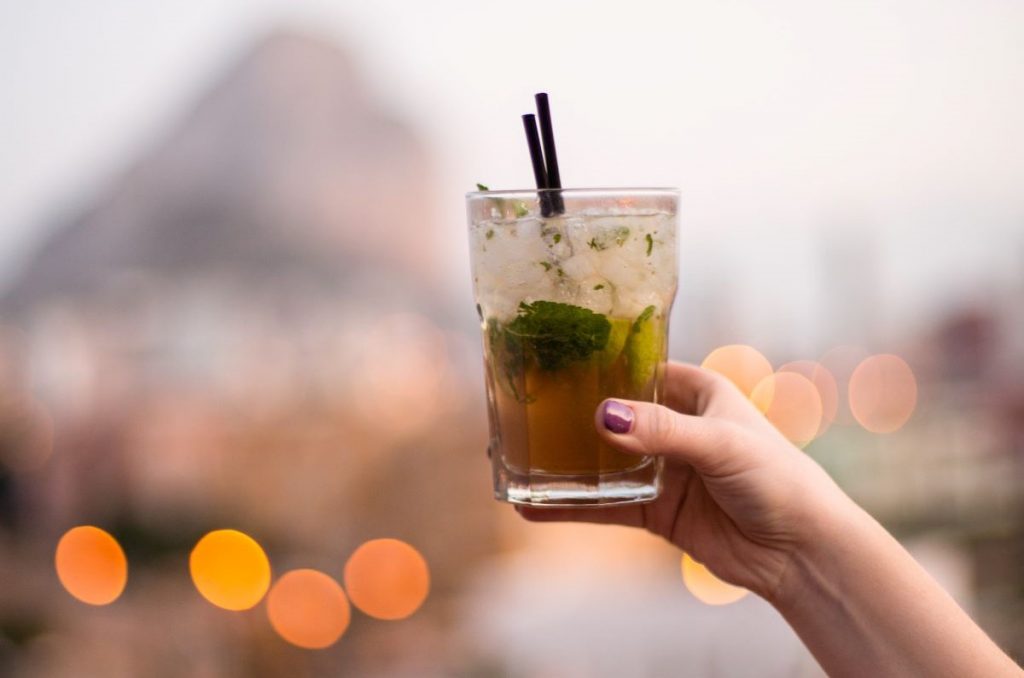 A person with purple nails holds a mint cocktail, overlooking Cape Town’s city lights and Table Mountain at sunset.