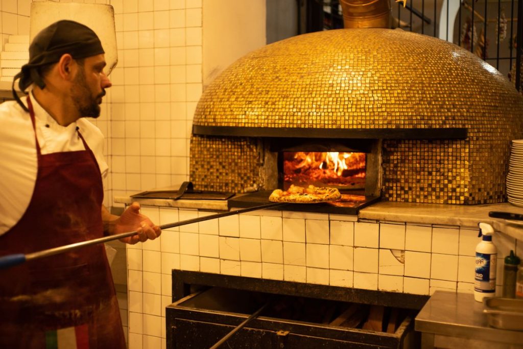 A chef at Pizzeria Da Michele in Naples slides pizza into a golden, wood-fired oven as bright flames flicker inside.