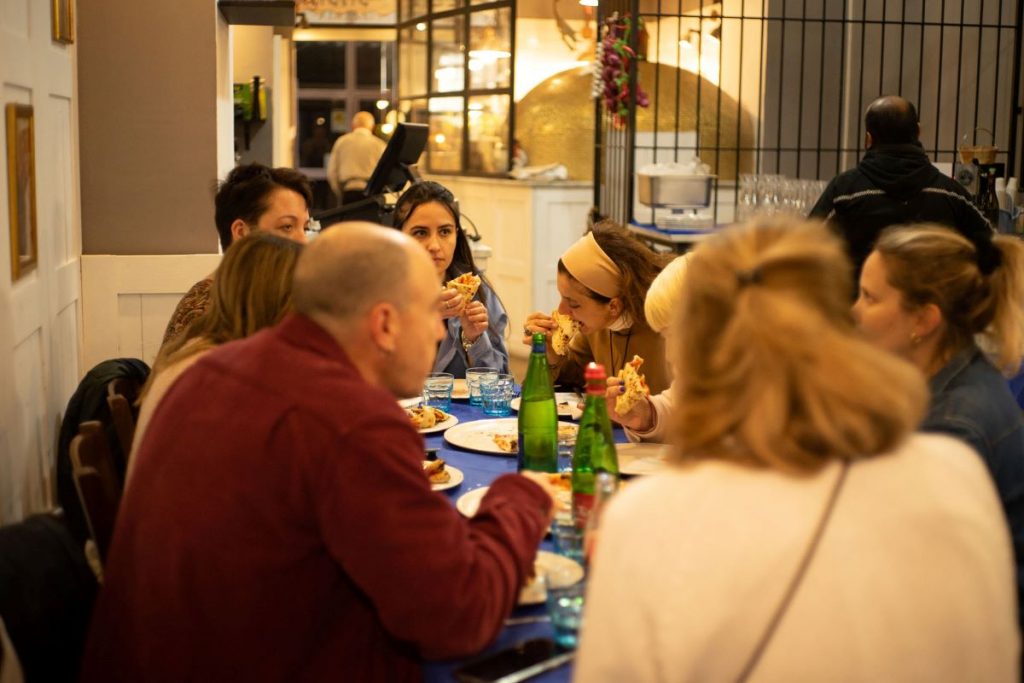Five friends share pizzas and laugh at a wooden table in Da Michele restaurant, Naples.