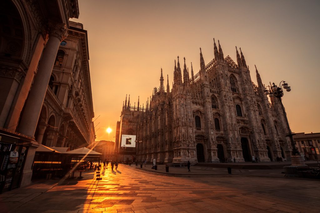 The sun sets behind Milan Cathedral in Italy, casting golden light across its detailed Gothic facade and the peaceful, empty square.