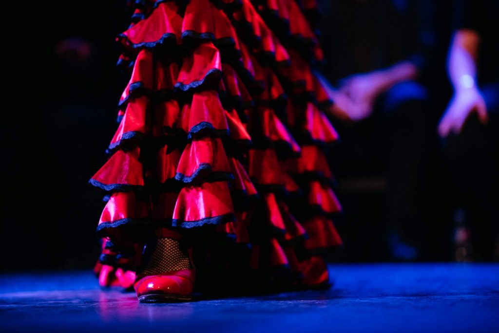A flamenco dancer performs on stage in a ruffled red and black dress, red shoes highlighted by spotlight, with musicians behind.
