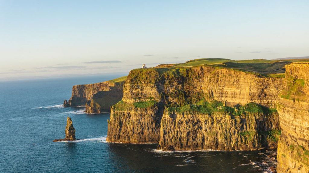 Sunlit cliffs at Ireland’s Cliffs of Moher overlook a blue ocean, with O’Brien’s Tower visible and waves hitting the rocks below.
