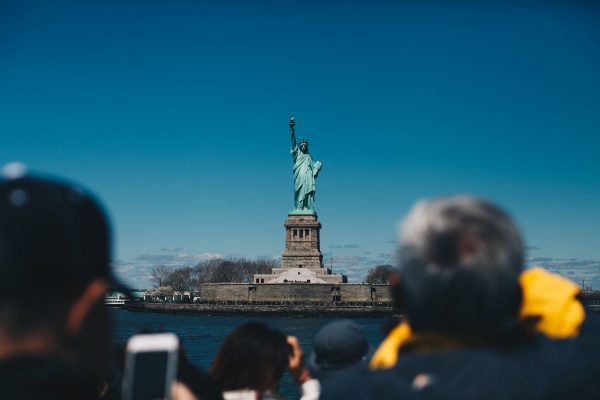 Visitors on a ferry point cameras toward the Statue of Liberty on Liberty Island, framed by a clear blue sky.