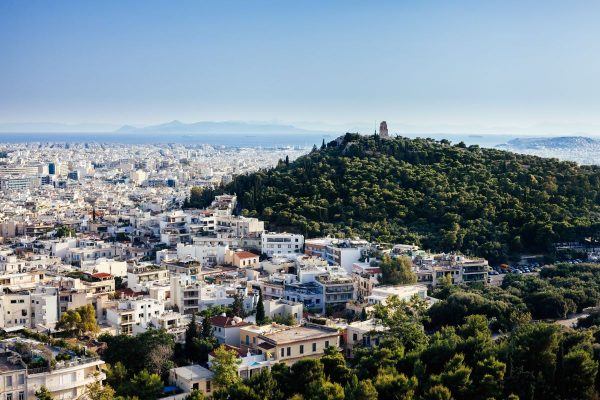 White buildings surround the tree-covered Lycabettus Hill in Athens, Greece, with the Chapel of St. George atop under clear skies.