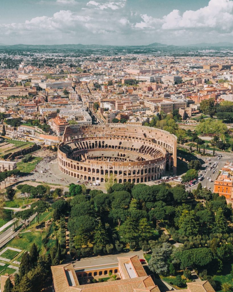 The Colosseum in Rome stands amidst city buildings, roads, green trees, and nearby restaurants under a partly cloudy sky.