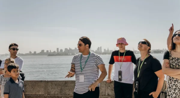 Several adults and children in sunglasses and casual clothes stand beside the Statue of Liberty ferry, with Manhattan skyline behind them.
