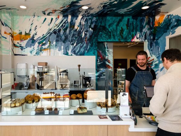 Man ordering baked goods from a cashier with a speckled mural behind him.