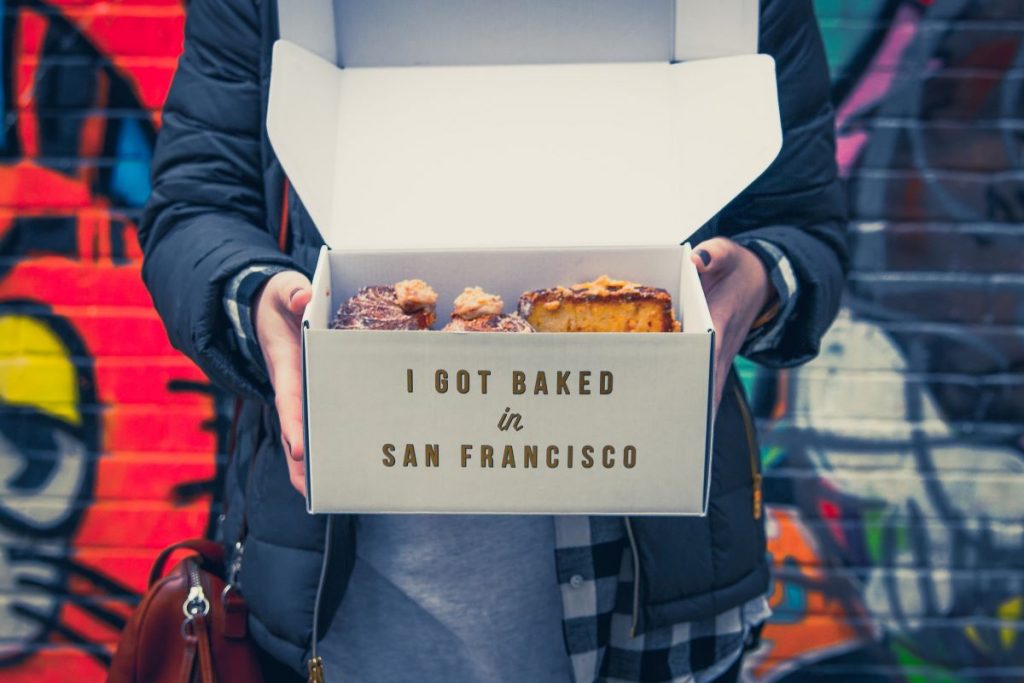 A person holds an open white bakery box of assorted pastries in front of a colorful graffiti wall in San Francisco.