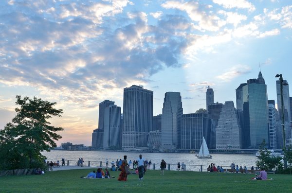 Several people lounge on green grass in Brooklyn Bridge Park, watching a sailboat drift along the East River at sunset.