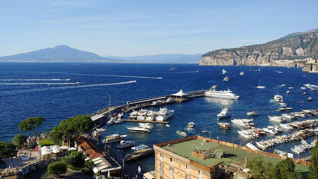 Numerous boats are docked at a marina in Sorrento, Italy; clear blue water, distant mountains, and waterfront buildings enhance the scene.