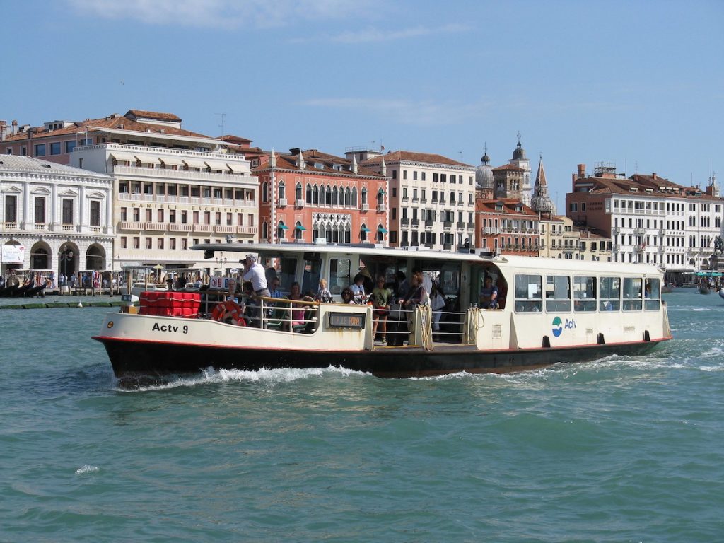 A Venice vaporetto carries passengers along the Grand Canal past colorful historic buildings and a bell tower on a sunny day.