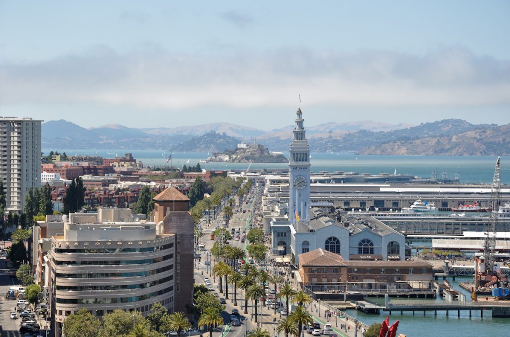 San Francisco’s waterfront shows the Ferry Building’s clock tower, palm-lined streets, and Alcatraz Island in the bay with surrounding hills.