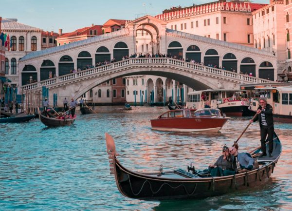 A gondolier rows a gondola with passengers along Venice’s Grand Canal, passing beneath the iconic Rialto Bridge and colorful buildings.